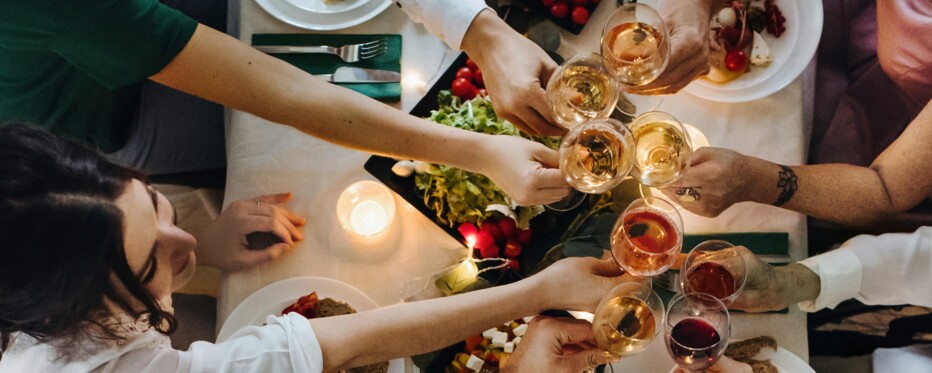 hands holding wine glasses reach over a dinner table to cheer each other