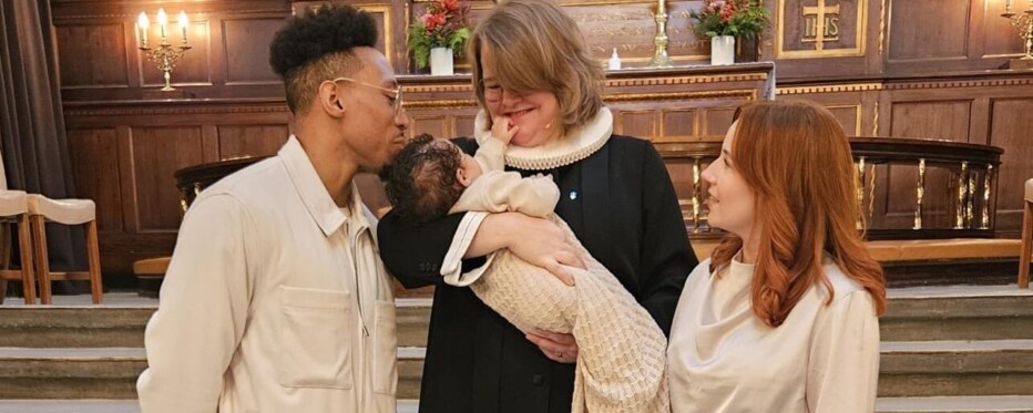 A new father and mother stand on either side of the Danish pastor who holds a newly baptised baby in her arms.